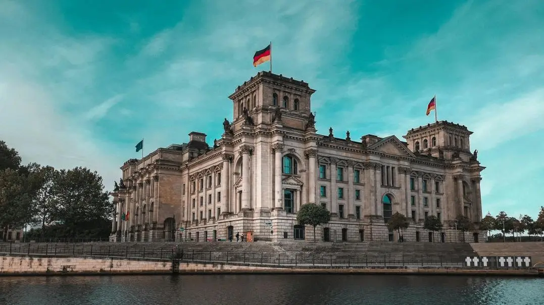 Daytime portrait of the Reichstag building in Berlin, capturing the detailed stonework, windows, and German flag on a pole in front.