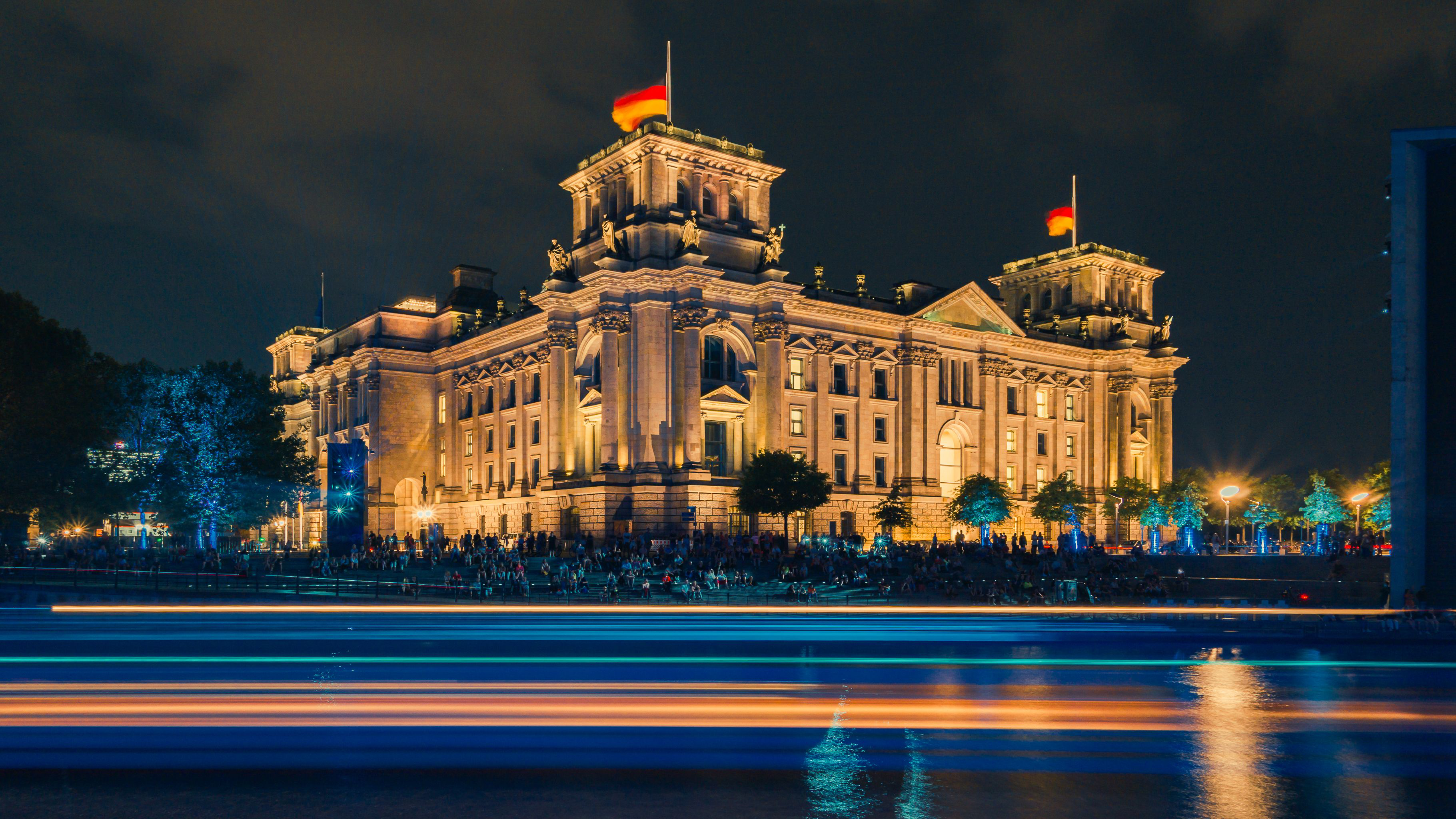 Nighttime view of the Reichstag building illuminated with golden lights, reflecting in the river in front, with people gathered along the embankment and German flags on top.