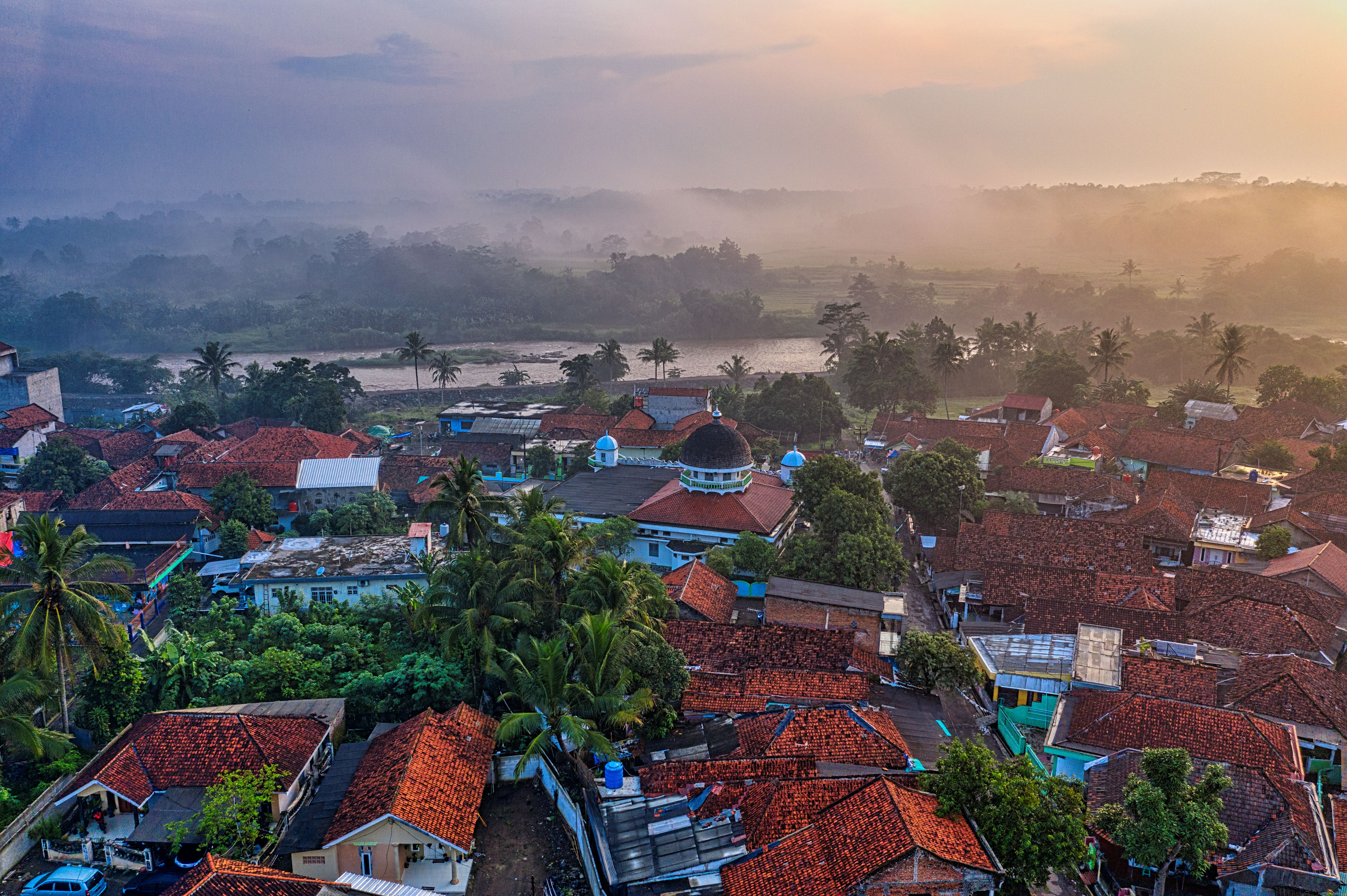 An Indonesian village at dawn with red-tiled roofs and a mosque surrounded by mist.